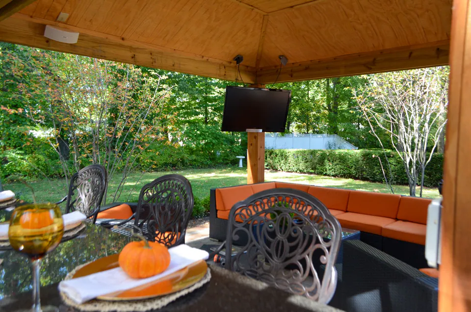 Close dining table view with pumpkin settings, outdoor television, orange lounge bench, wood ceiling, and garden backdrop.